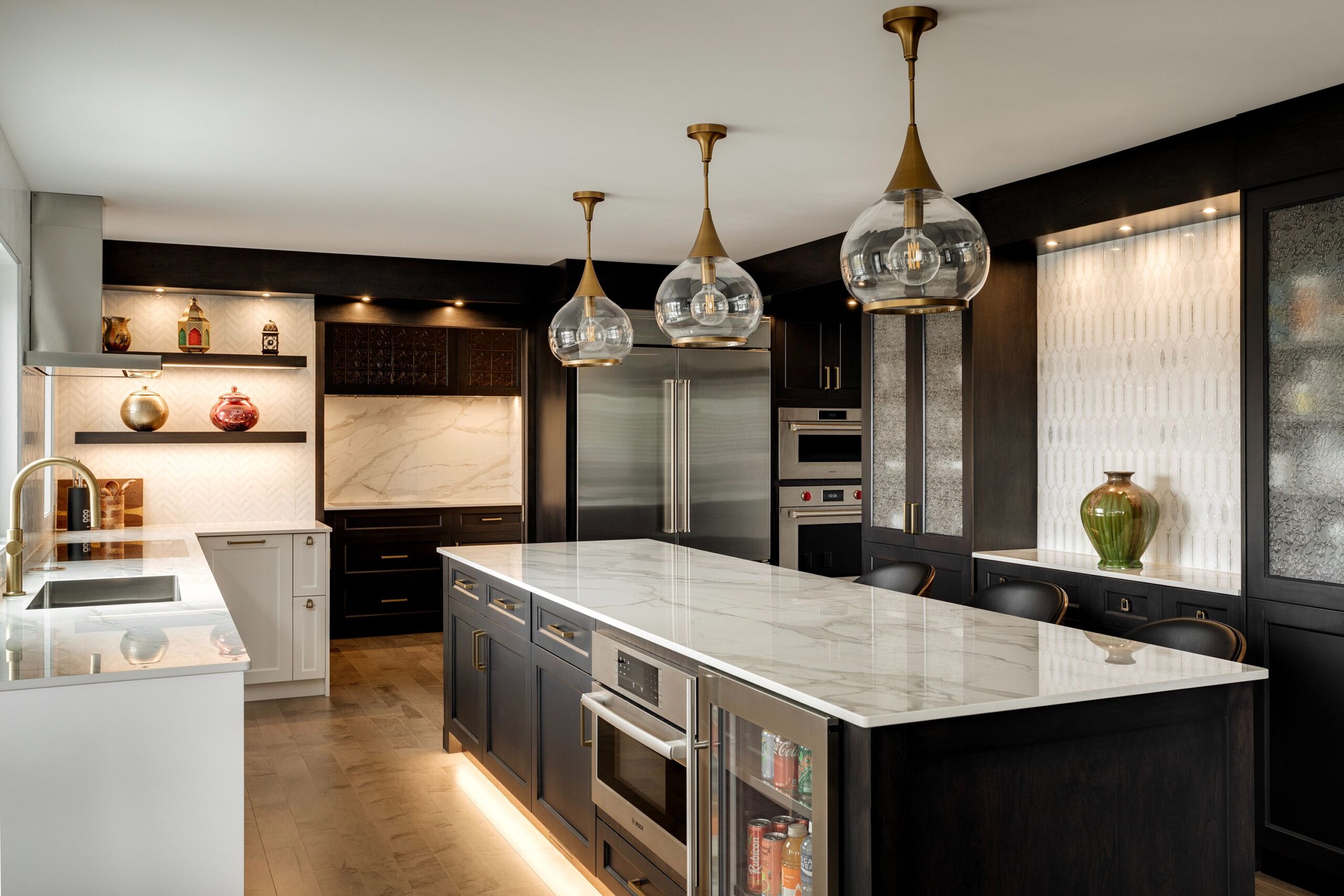 A modern kitchen with dark cabinets, a large marble island, built-in appliances, glass pendant lights, open shelves with decor, and under-cabinet lighting on a wooden floor.