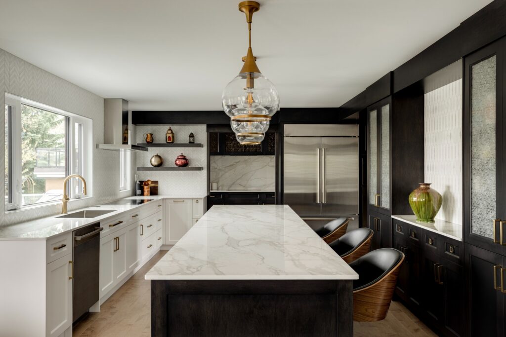 Modern kitchen with a large marble island, black and white cabinetry, gold fixtures, built-in appliances, decorative vases, and three black chairs. Large windows provide natural light.