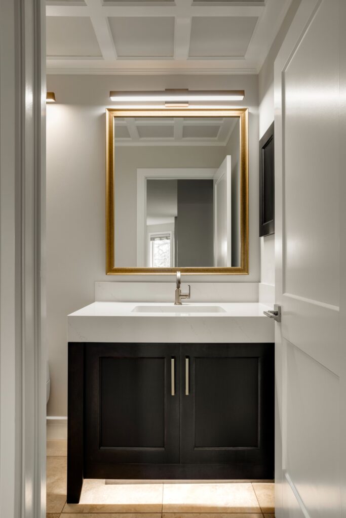 A modern bathroom features a black vanity with a white countertop, a rectangular sink, a gold-framed mirror, and a wall light above. The room has white walls, a coffered ceiling, and a partially open door.