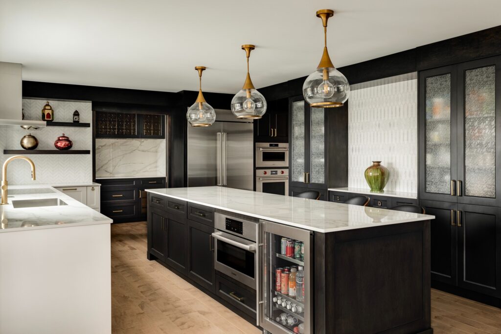 Modern kitchen with dark cabinets, a large white marble island, built-in appliances, three glass pendant lights, and decorative vases. The island features a glass-front beverage fridge and seating area, with wood flooring throughout.