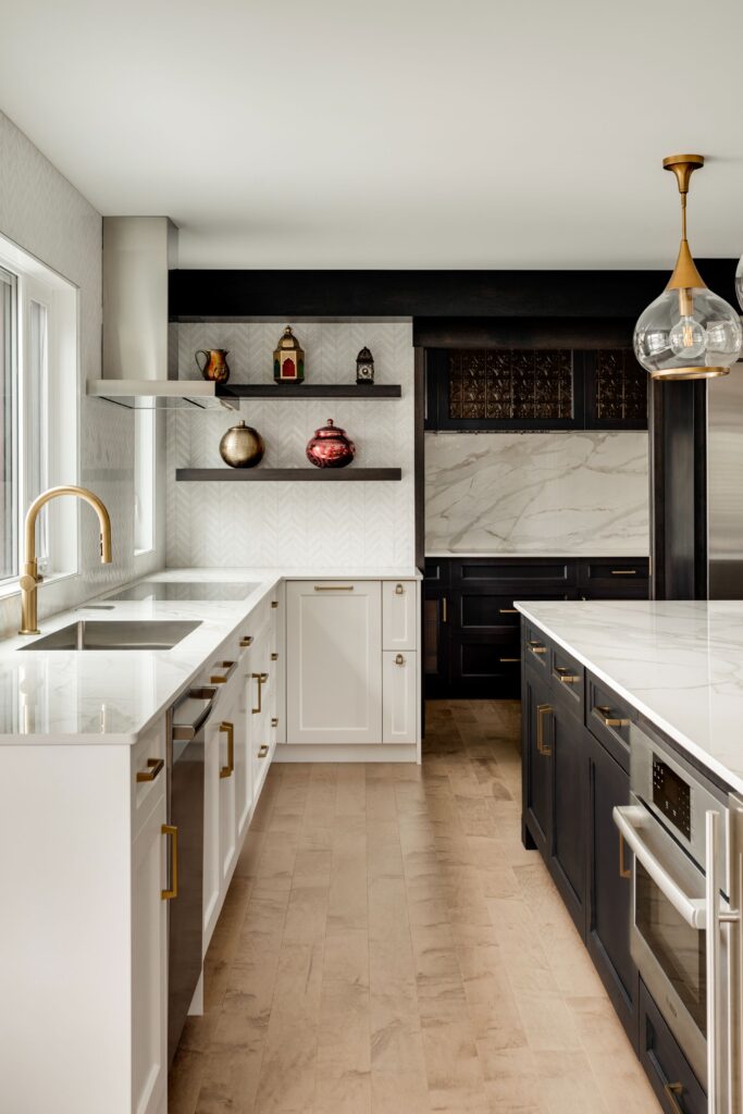 Modern kitchen with white and dark cabinets, gold hardware, marble countertops, open shelves with decorative items, a gold faucet, built-in appliances, and light wood floors. Large window provides natural light.