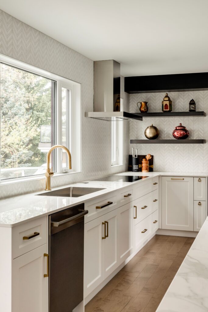 Modern kitchen with white cabinets, gold handles, marble countertops, a large window, stainless steel appliances, and open black shelves displaying decorative items against a light patterned backsplash.