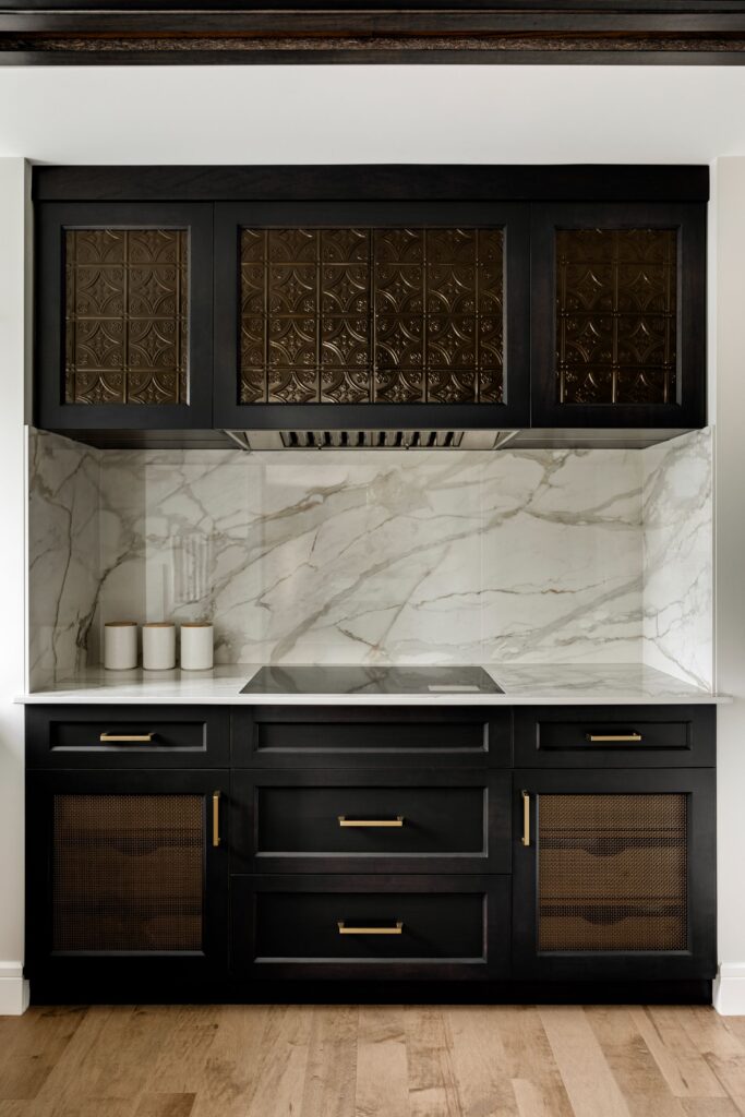 Modern kitchen with dark wood cabinets, gold handles, a marble backsplash, and three white canisters on the counter. The upper cabinets have decorative glass panels, and the floor is light wood.