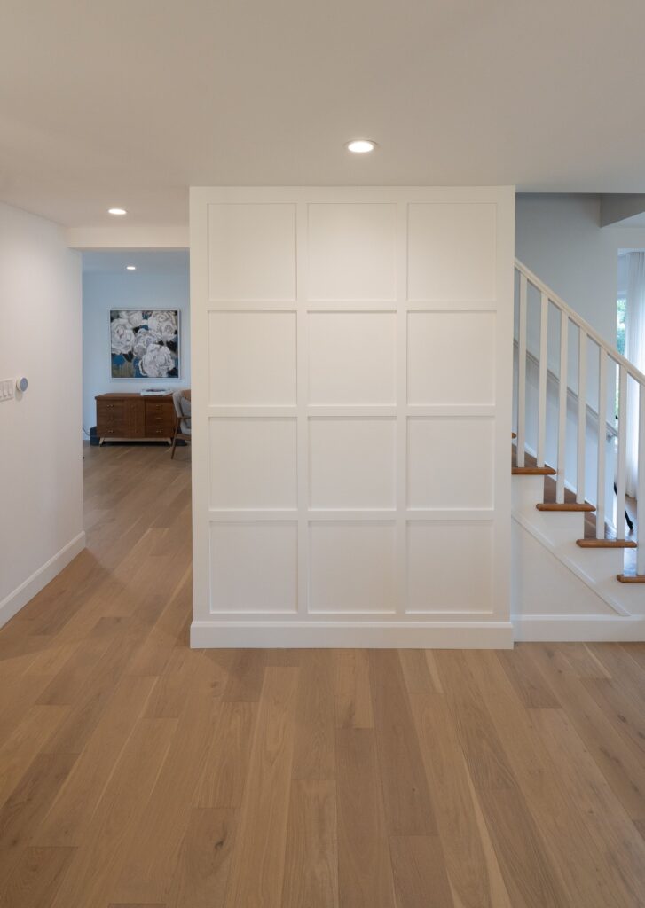 A modern interior with light wood floors, a white paneled accent wall, and a staircase with white railings. In the background, a wooden sideboard and large floral artwork are visible. The space is brightly lit.
