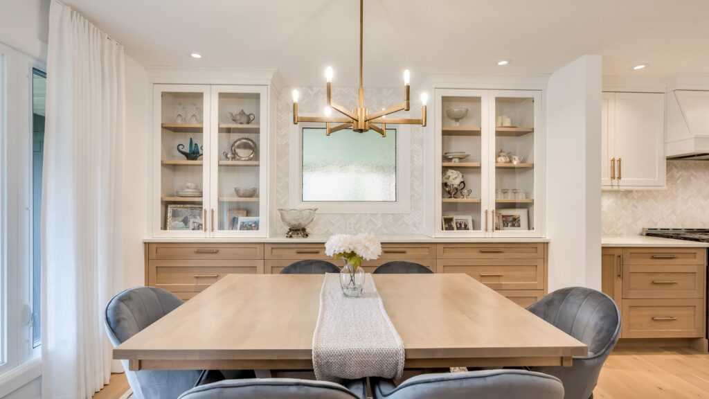 A modern dining room with a light wood table, six gray cushioned chairs, a vase of white flowers, and a gold chandelier. Built-in cabinets with glass doors and decor items line the back wall. White curtains cover a large window.