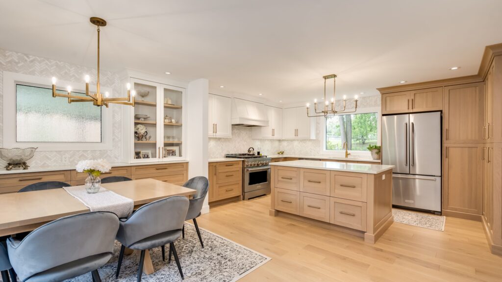 Modern kitchen and dining area featuring light wood floors, beige cabinetry, stainless steel appliances, two chandeliers, a center island, built-in shelves, and a dining table with six gray upholstered chairs.
