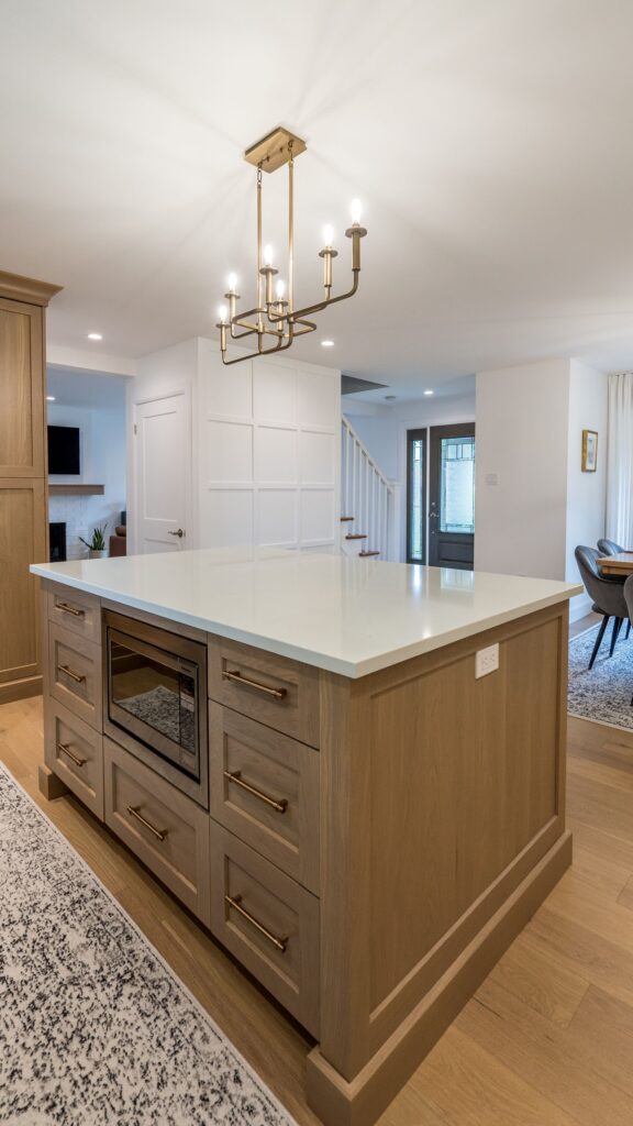 A modern kitchen with a large wooden island featuring drawers, a built-in microwave, and a white countertop. Overhead, a gold chandelier hangs. The open floor plan reveals stairs, a dining area, and a glimpse of the living room.