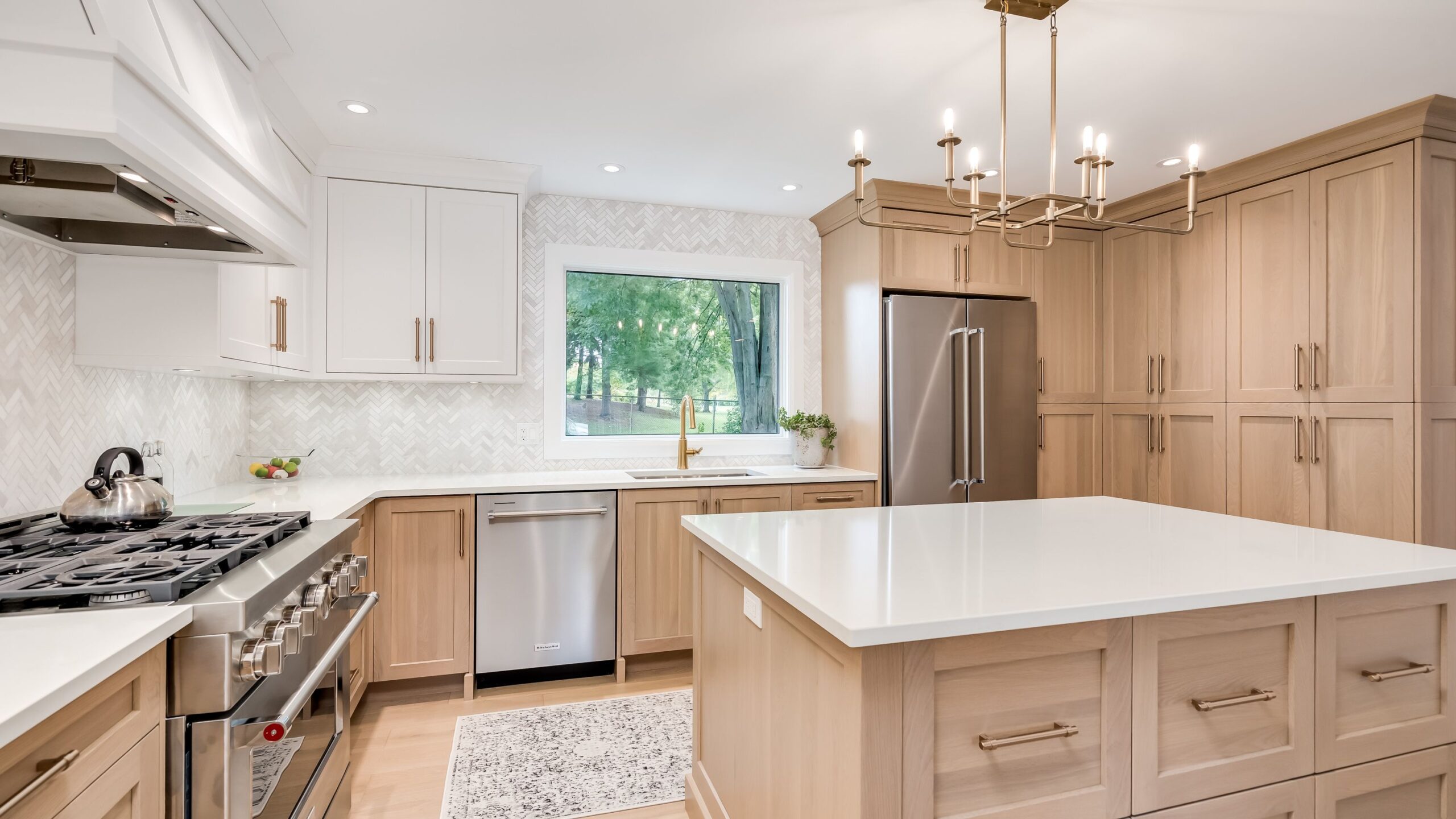 Modern kitchen with light wood cabinets, white countertops, stainless steel appliances, a large center island, a window with a view of greenery, and a gold chandelier hanging from the ceiling.