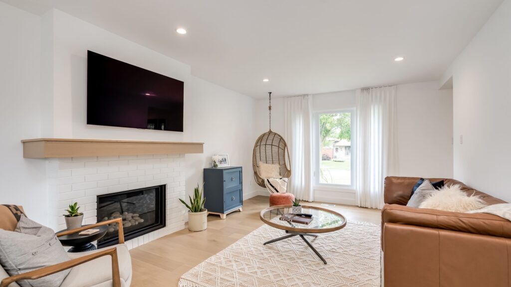 Bright living room with a mounted TV above a fireplace, a brown leather sofa, round glass coffee table, woven hanging chair by a window, armchair, blue cabinet, and light wood flooring.