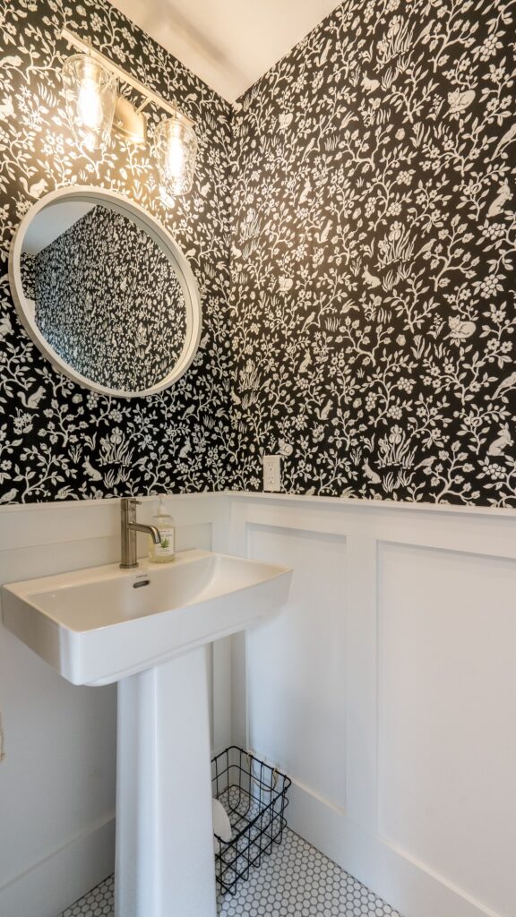 A small bathroom with black and white floral wallpaper, an oval mirror, a white pedestal sink with a silver faucet, two wall lights, white wainscoting, hexagonal tile floor, and a small black wire basket.