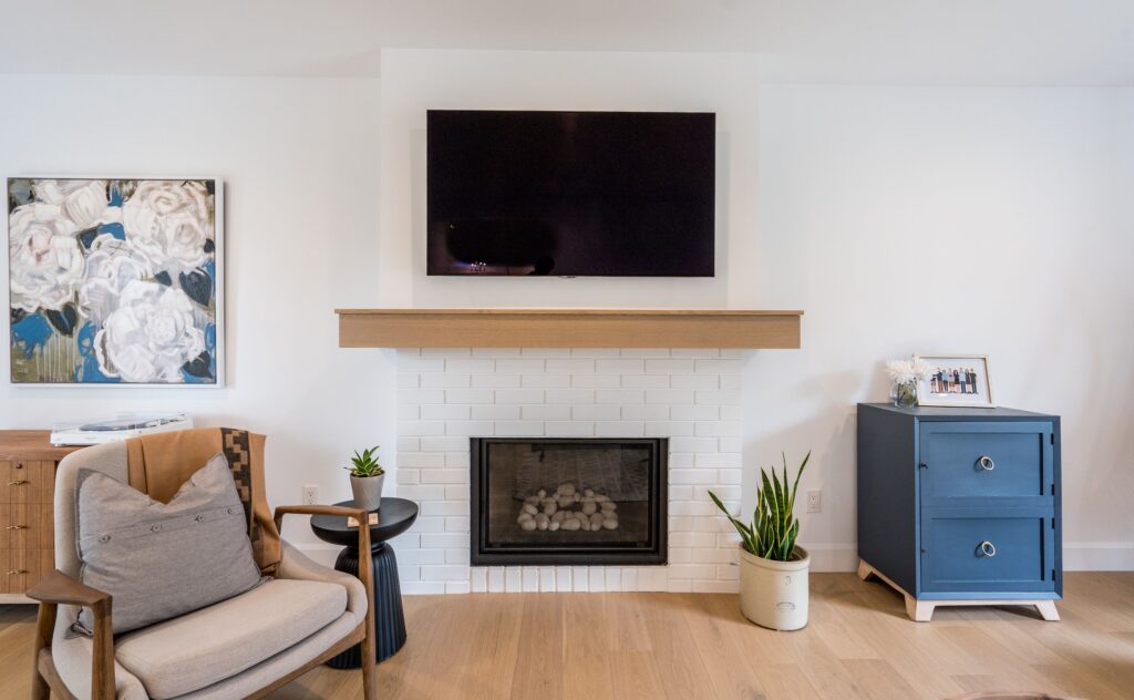 A modern living room with a white brick fireplace, wooden mantel, and TV above. A blue cabinet with a framed photo, a potted plant, a beige armchair, a small side table, and floral wall art decorate the space.