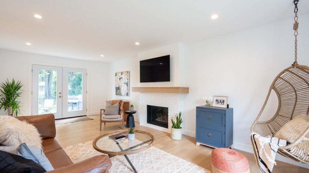 Bright living room with a brown leather sofa, round glass coffee table, hanging wicker chair, blue cabinet, fireplace with TV above, and large windows letting in natural light.