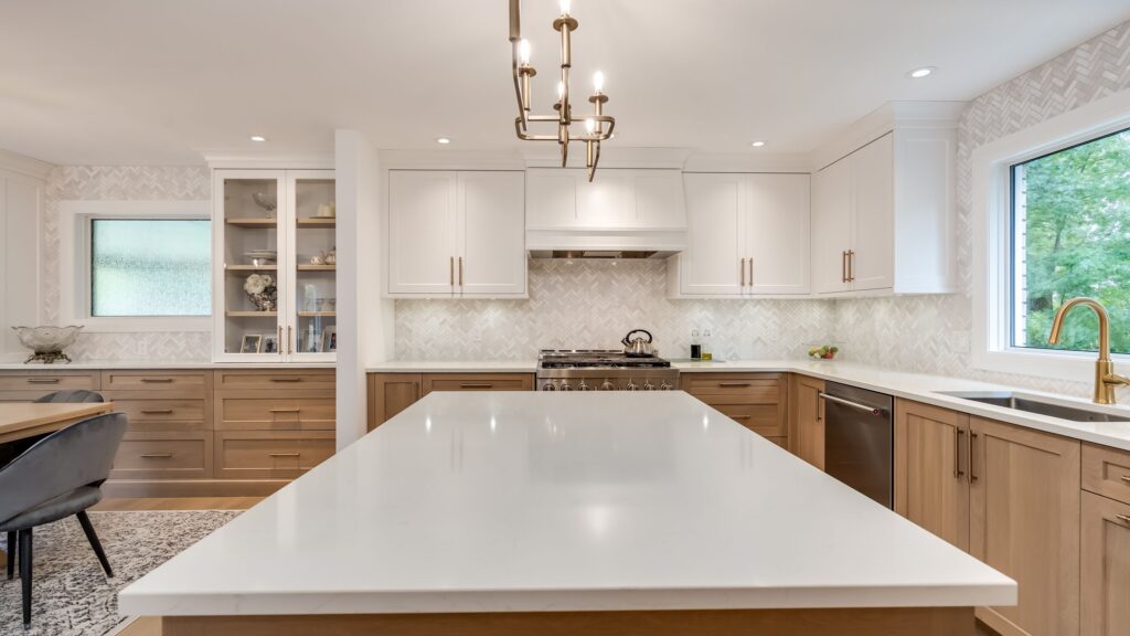 Modern kitchen with a large white island, wooden lower cabinets, white upper cabinets, stainless steel appliances, gold faucet, pendant lights, and large windows letting in natural light.