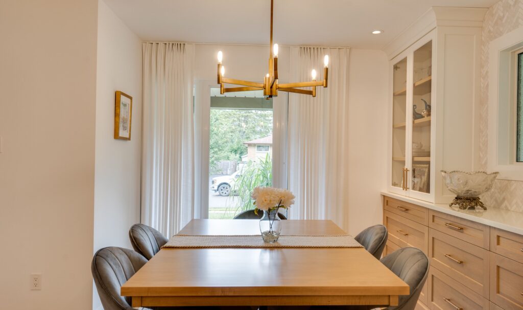 A modern dining room with a wooden table, four gray chairs, a gold chandelier, white curtains, and built-in cabinets. A vase of white flowers sits on the table, and glass doors reveal a view of a green backyard.