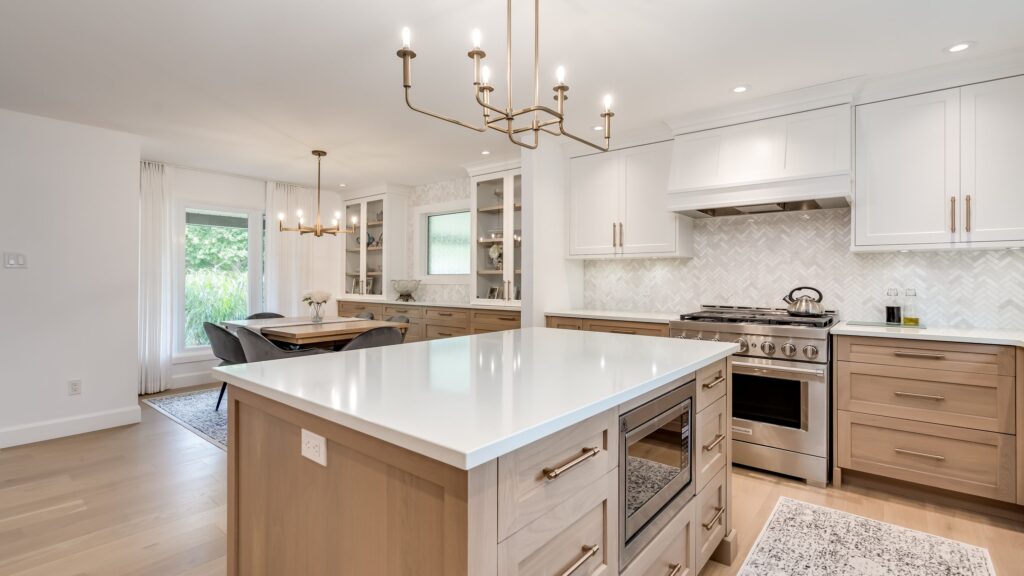 Modern kitchen with light wood cabinets, white countertops, stainless steel appliances, a large island with built-in microwave, and two gold chandeliers. There is a dining area with a window in the background.