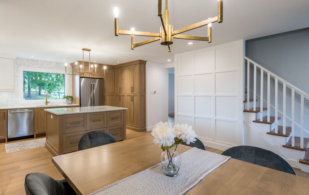 Modern kitchen and dining area with wood cabinets, island, stainless steel appliances, chandelier, and a vase of white flowers on the dining table. Stairs with wooden steps are visible on the right.