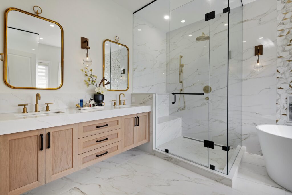 Modern bathroom with double sink vanity, gold fixtures, large glass-enclosed shower, marble floors and walls, wall-mounted mirrors, and a white freestanding bathtub. Decor includes an orchid and pendant lights.