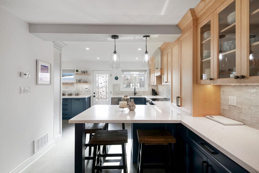 Modern kitchen with light wood cabinets, navy blue lower cabinets, a white quartz countertop island, three wooden stools, pendant lights, and a windowed door in the background. The space is bright and tidy.