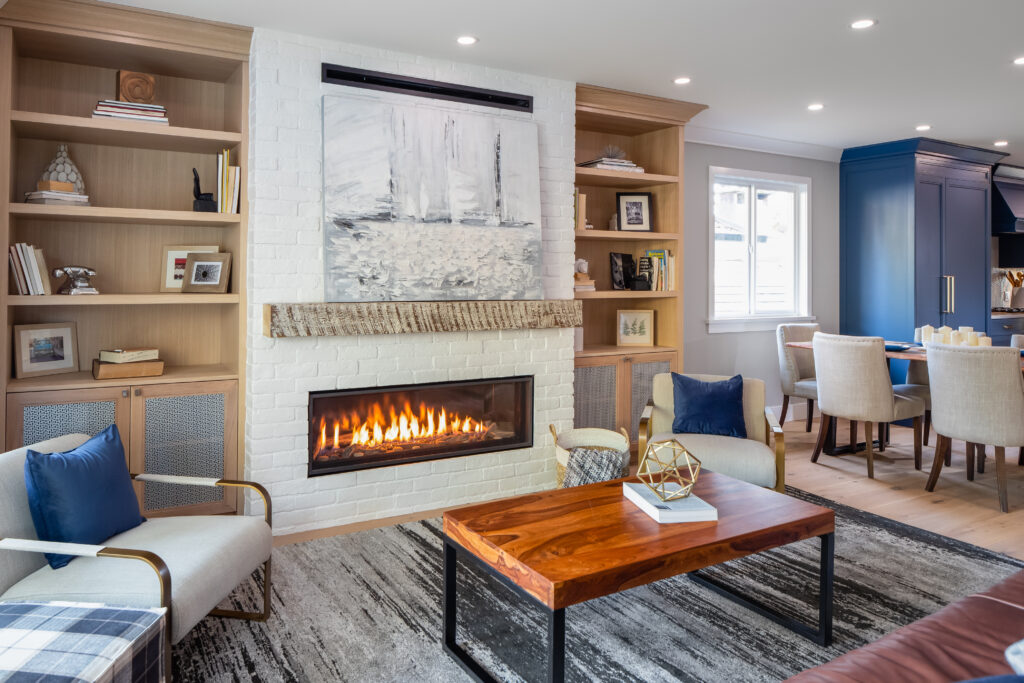A modern living room features a white brick fireplace with a painting above it, flanked by built-in wooden shelves. A wood coffee table, two armchairs, and a rug complete the cozy, well-lit space next to a dining area.