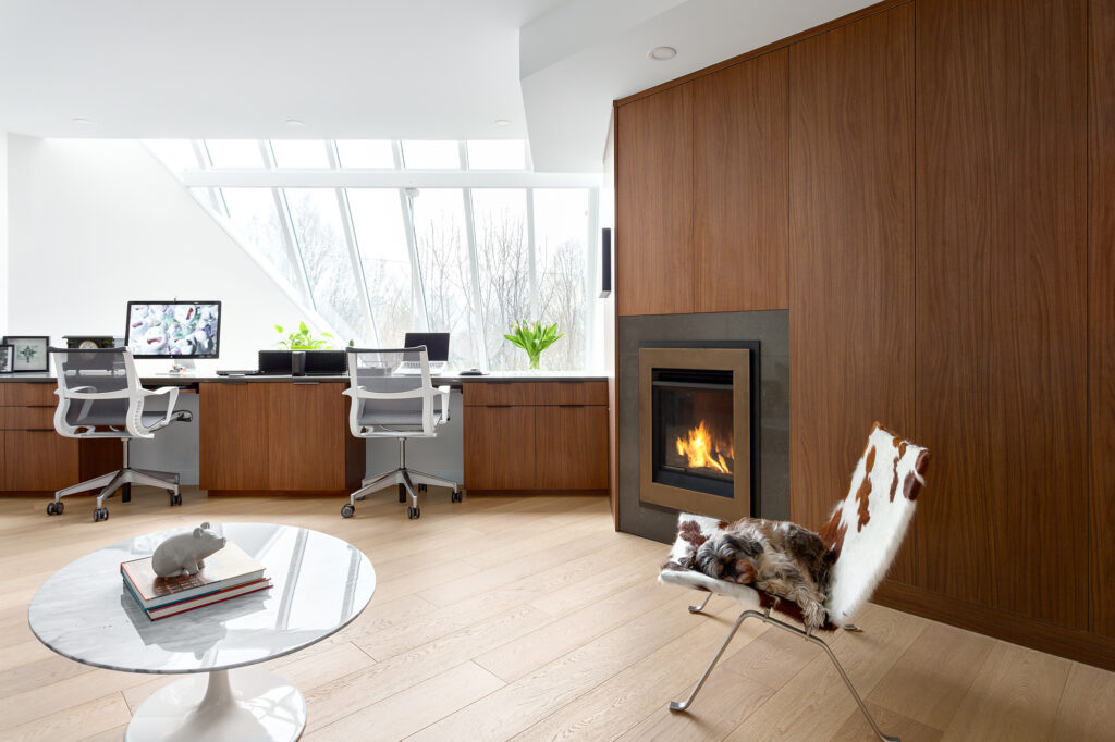 Modern home office with two desks, white chairs, and computers by large windows; a cozy fireplace, a cowhide chair with a small dog resting, and a round table with books and a small sculpture. Natural light fills the space.