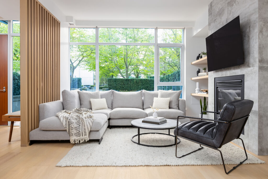 A modern living room with a light gray sectional sofa, a black leather chair, a round coffee table, a soft rug, and a wall-mounted TV above a fireplace. Large windows let in natural light and show green trees outside.