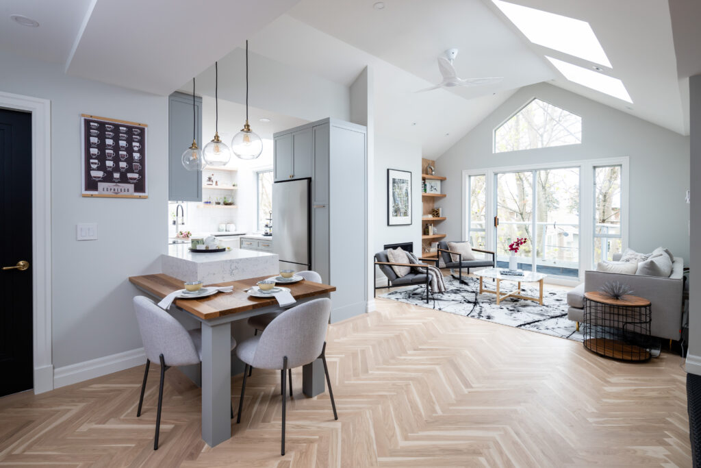 Modern open-concept kitchen and living room with natural light, light wood herringbone floors, white cabinets, a small dining table set for two, and a cozy sitting area near large windows and glass doors.