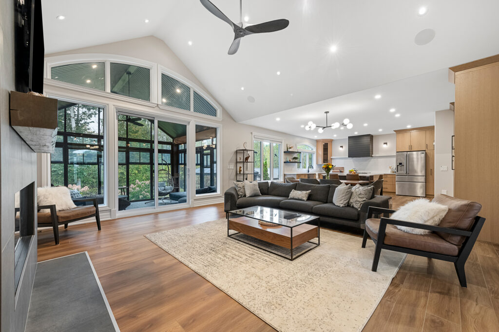 Open-concept living room and kitchen with vaulted ceilings, modern furnishings, large windows, sliding doors to a screened patio, wooden floors, and neutral decor, creating a bright, spacious, and inviting space.