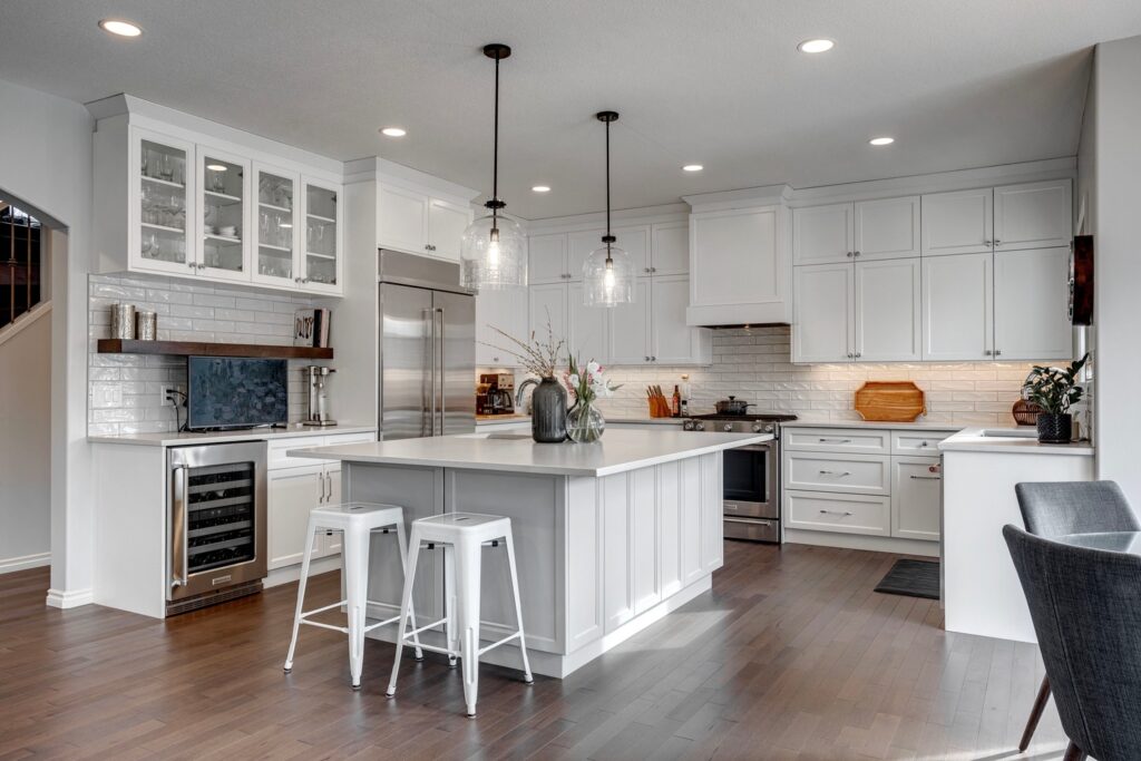 Modern kitchen with white cabinets, stainless steel appliances, a large island with two white stools, pendant lights, dark wood floors, and decorative accents on the island and counters.