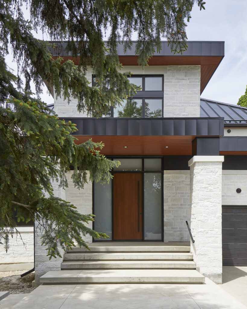 A modern two-story house with light stone exterior, large black-framed windows, a wooden front door, and steps leading to the entrance, partially framed by green pine tree branches.