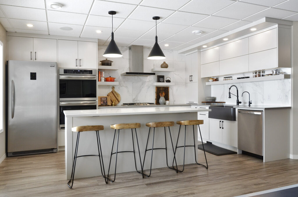 Modern kitchen with white cabinets, stainless steel appliances, a kitchen island with white countertop, three wooden bar stools, pendant lights, and a farmhouse sink. Light wood flooring and bright lighting complete the space.