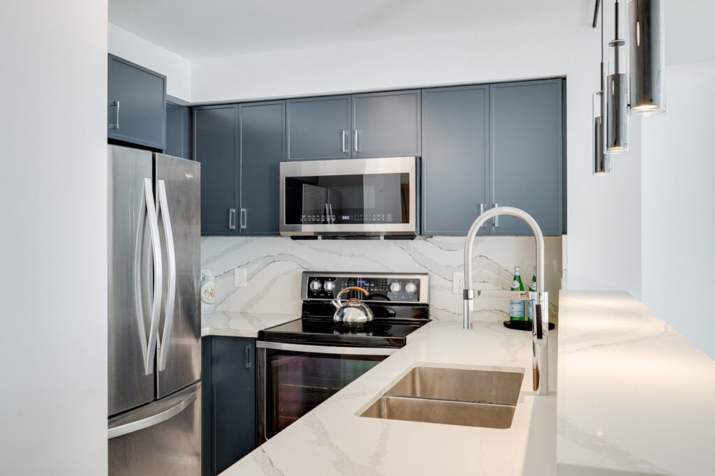 Modern kitchen with dark blue cabinets, stainless steel appliances, a double sink, electric stove, microwave, and marble-patterned countertops and backsplash. Three pendant lights hang over the white counter.