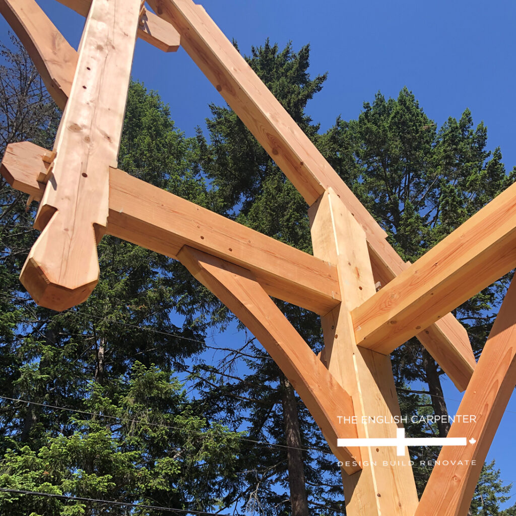 Wooden timber frame structure under construction, viewed from below against a clear blue sky and tall green trees. The logo The English Carpenter - Design. Build. Renovate. appears in the lower right corner.