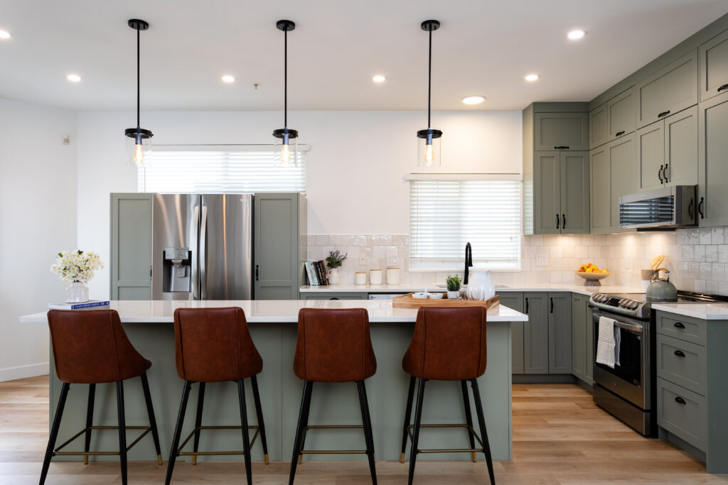 Modern kitchen with sage green cabinets, stainless steel appliances, a white island with four brown leather bar stools, pendant lights, and wooden flooring. Windows provide natural light and fresh flowers are on the counter.