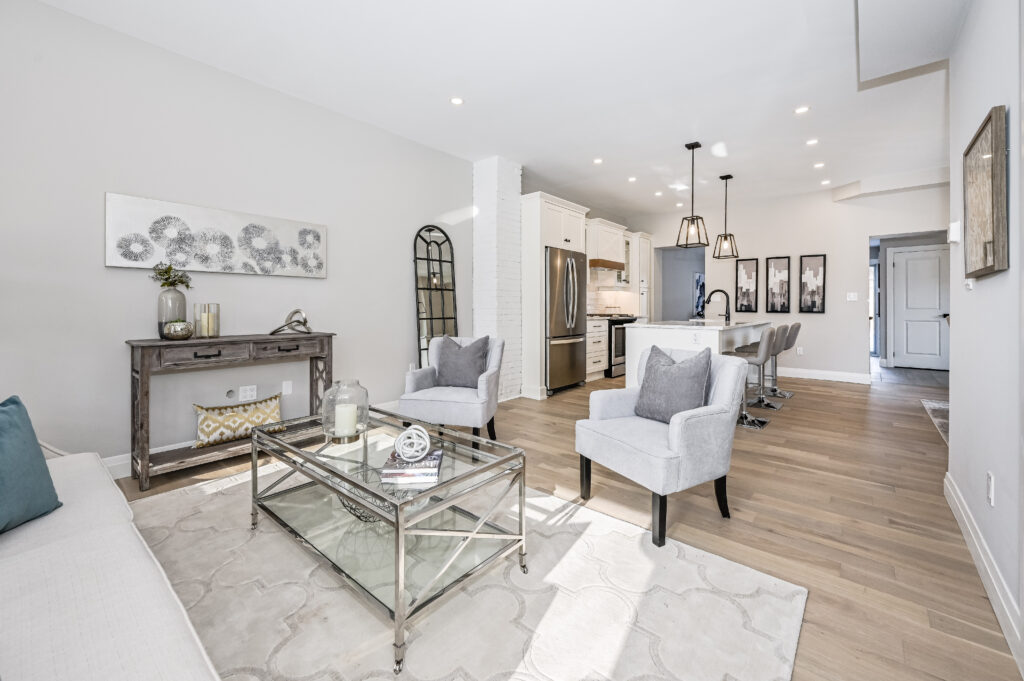 Bright open-concept living and kitchen area with light wood floors, neutral furnishings, two gray armchairs, glass coffee table, kitchen island with barstools, and modern pendant lighting.