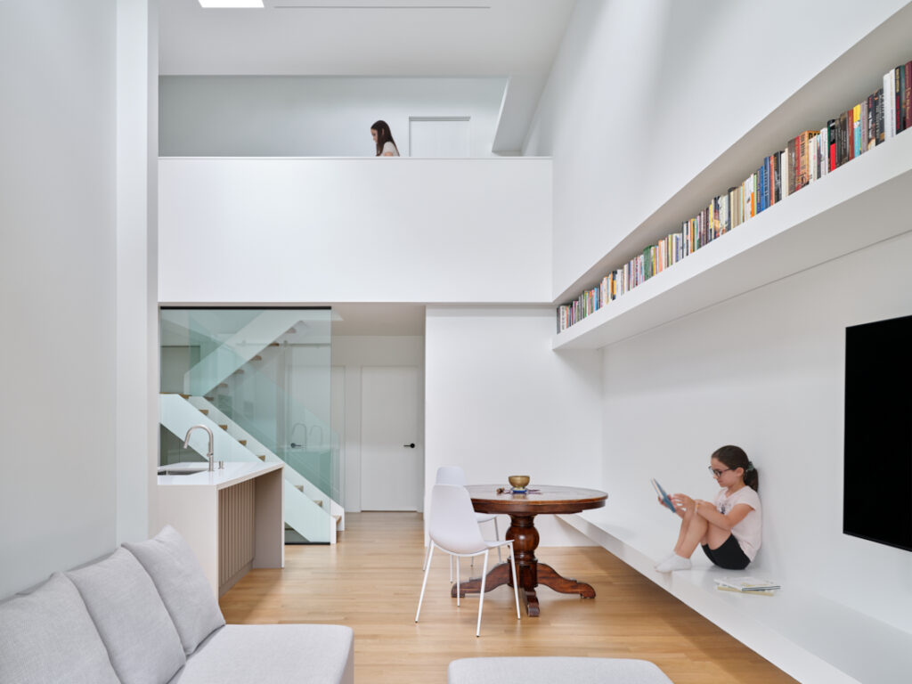 A modern, minimalist living space with white walls and light wood floors. A girl reads on a built-in bench below a long bookshelf, while another person walks on the upper level. A round table, chairs, and a sofa complete the room.