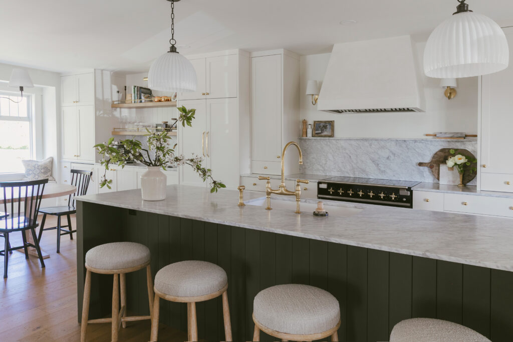 A modern kitchen with white cabinets, marble countertops, a large island with three cushioned stools, gold fixtures, hanging pendant lights, and a dining area with a wooden table and chairs by a window.