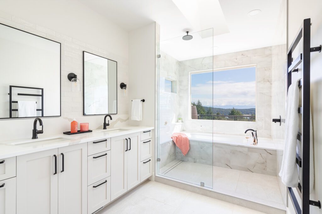 A modern, bright bathroom with white cabinets, double sinks, black fixtures, a glass shower, and a bathtub by a large window with a scenic view. A coral towel adds a pop of color.