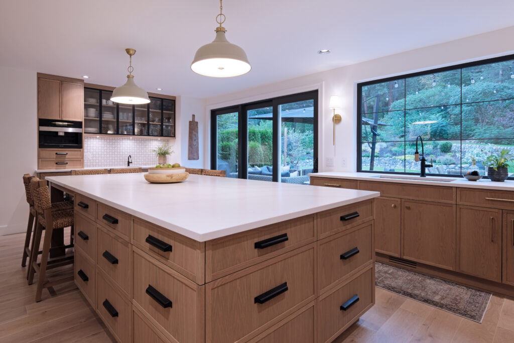 Modern kitchen with light wood cabinets, a large white island with black handles, pendant lights, bar stools, large windows, and glass doors opening to a lush outdoor area. The space feels bright and spacious.