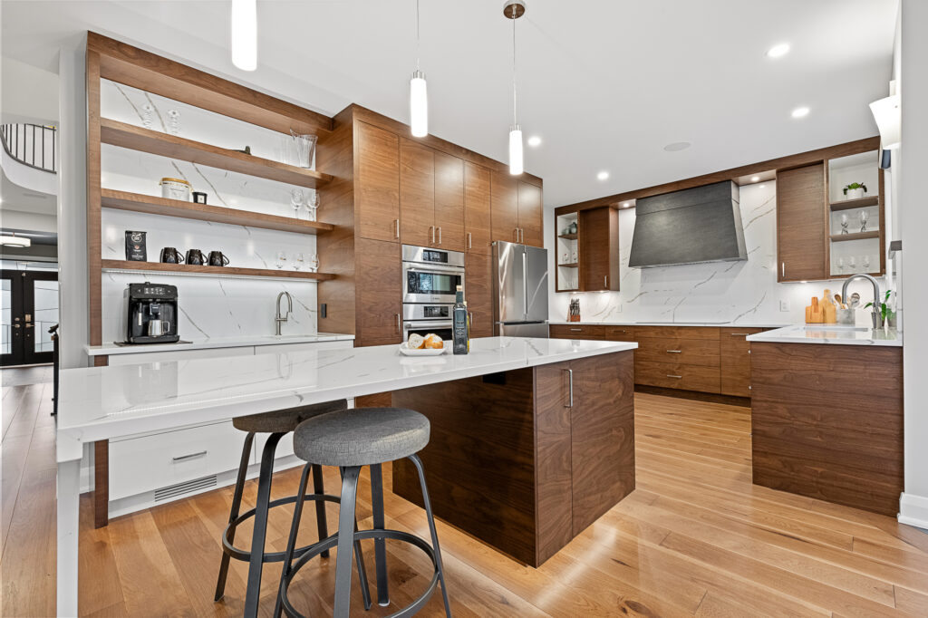 Modern kitchen with wooden cabinets, white countertops, built-in appliances, open shelves, pendant lights, and a large island with two stools. The space features light wood flooring and a bright, clean design.