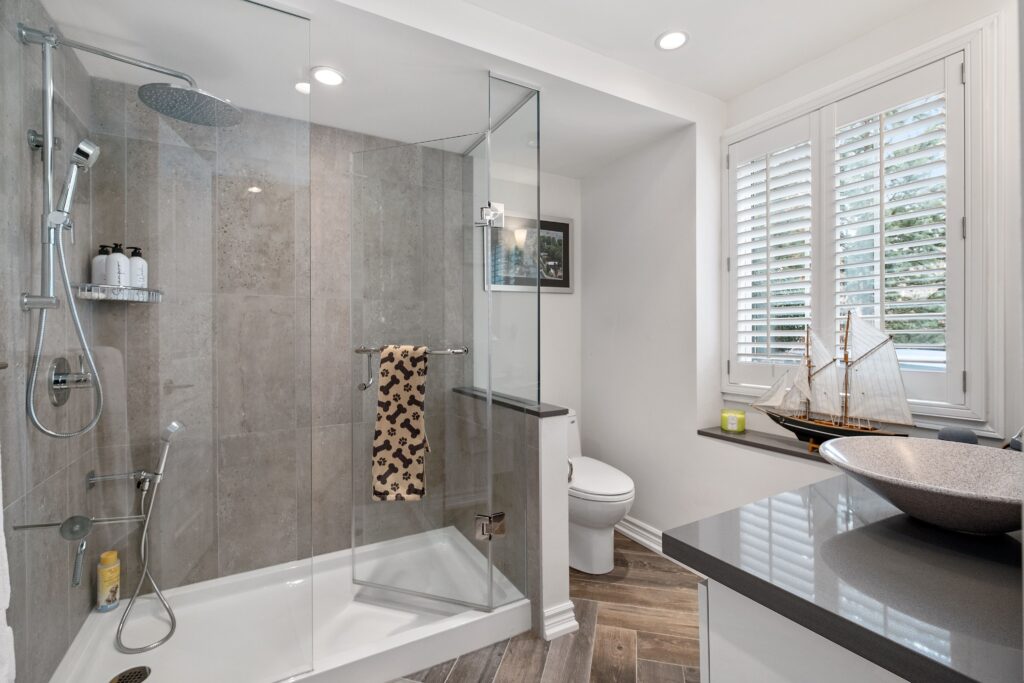 Modern bathroom with a glass shower enclosure, gray tiles, a rainfall showerhead, and a towel with a leopard print. There’s a toilet, a countertop with a vessel sink, and a window with white shutters. A model ship decorates the windowsill.