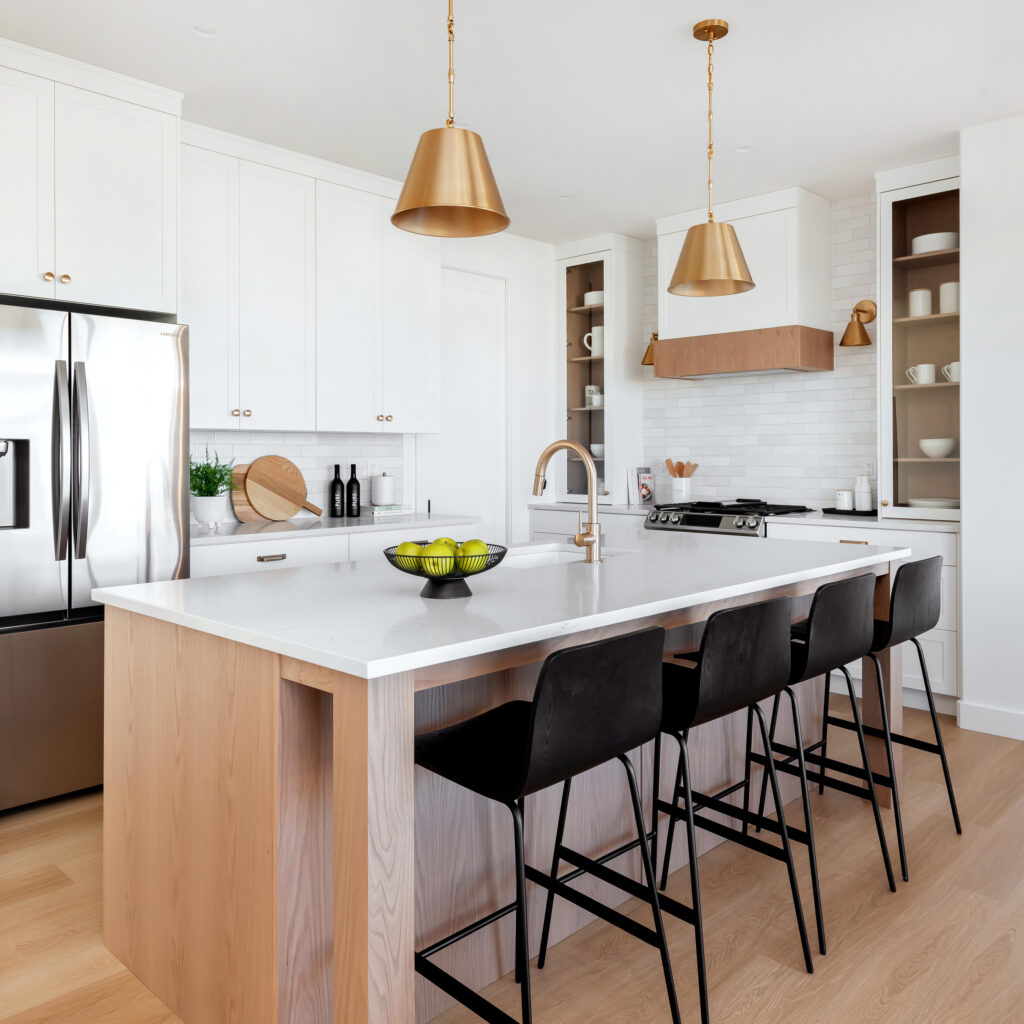 Modern kitchen with a large island featuring a white countertop, light wood base, four black barstools, stainless steel appliances, white cabinets, gold pendant lights, and decorative bowls and utensils.