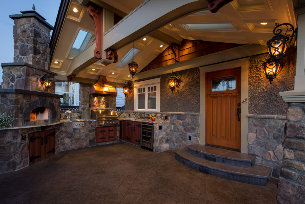 Outdoor kitchen with stone walls, built-in grill, hood, cabinets, wine fridge, and decorative lantern lights under a covered patio near a wooden door; the area is illuminated warmly in the evening.