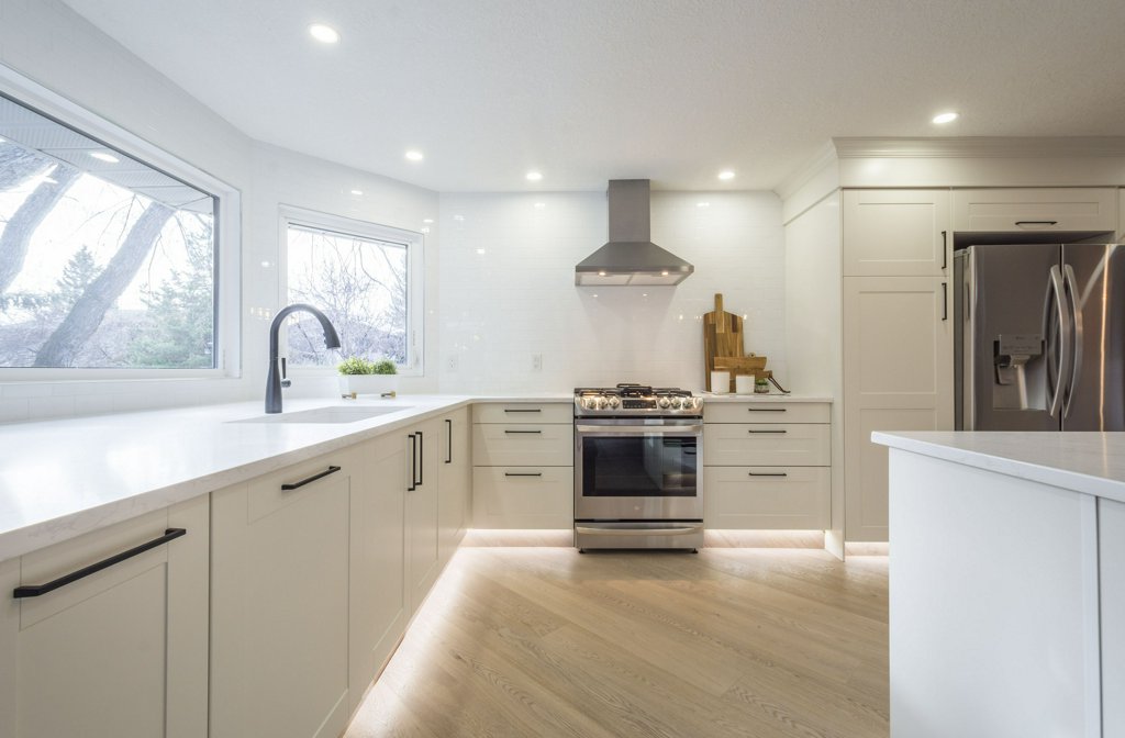 Bright, modern kitchen with white cabinets, stainless steel appliances, a gas stove with a hood, large windows, light wood floors, and minimalistic black handles. Natural light enhances the clean, spacious design.