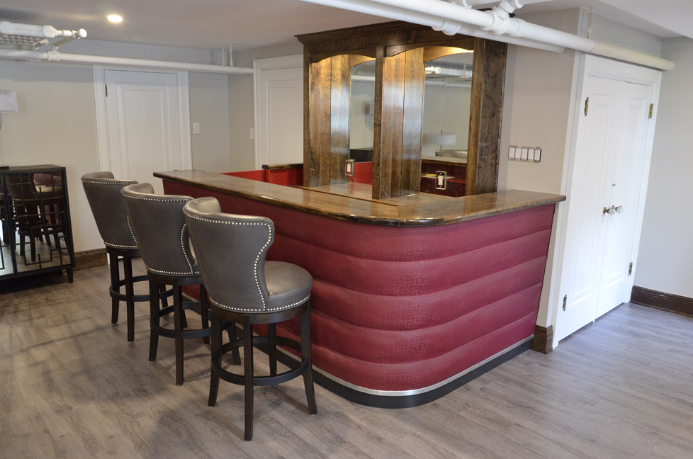 A stylish home bar with a curved red front, wooden countertop, and mirrored back shelves. Three gray upholstered bar stools with nailhead trim are lined up at the counter. The room has wood flooring and white doors.