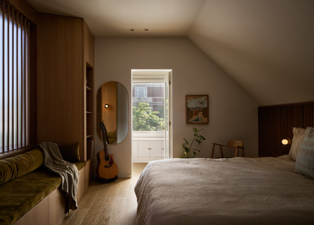 A cozy bedroom with wood accents, a large bed, a window seat, a guitar by an oval mirror, a plant, a chair, and a bright window showing trees and a brick building outside.
