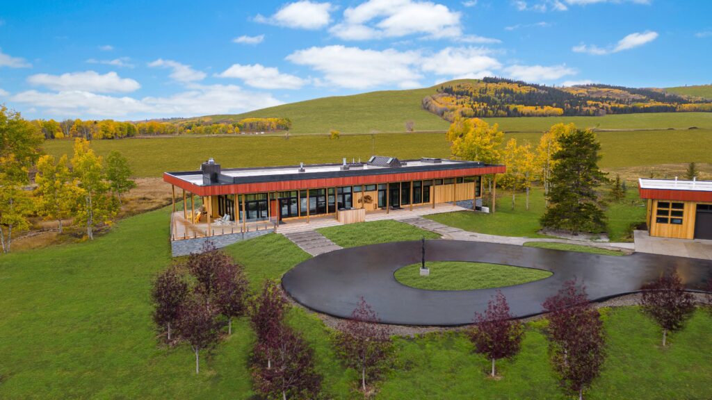 A modern, single-story building with large windows sits on a green lawn with a circular driveway, surrounded by trees and rolling hills under a blue sky with scattered clouds.