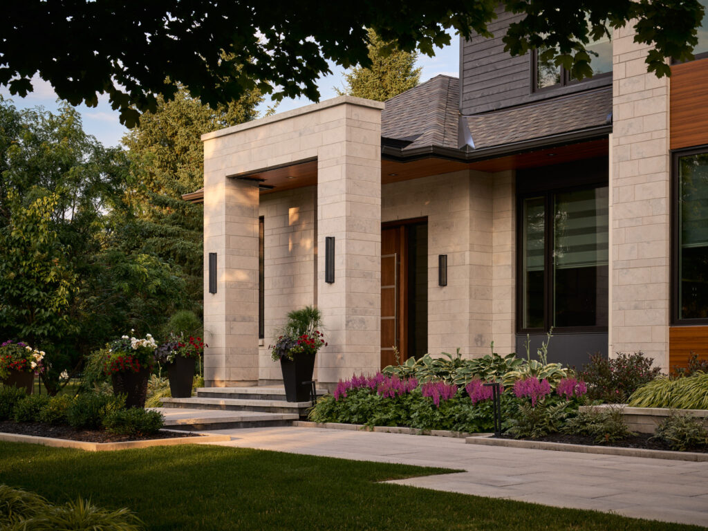 Modern house exterior with light stone walls, large windows, and wood accents. A landscaped garden with purple flowers, shrubs, and potted plants surrounds the entrance. Trees and green lawn are in the foreground.
