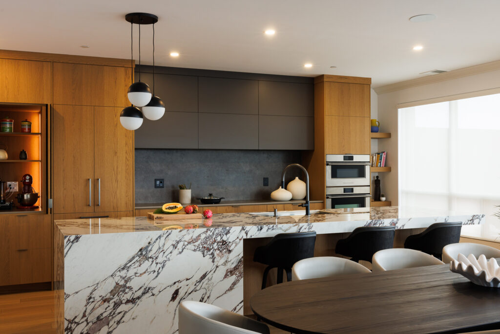 Modern kitchen with wooden cabinets, a marble island, pendant lights, built-in ovens, open shelving, and black and white chairs around a dining table. The countertop features fruit and decorative objects.