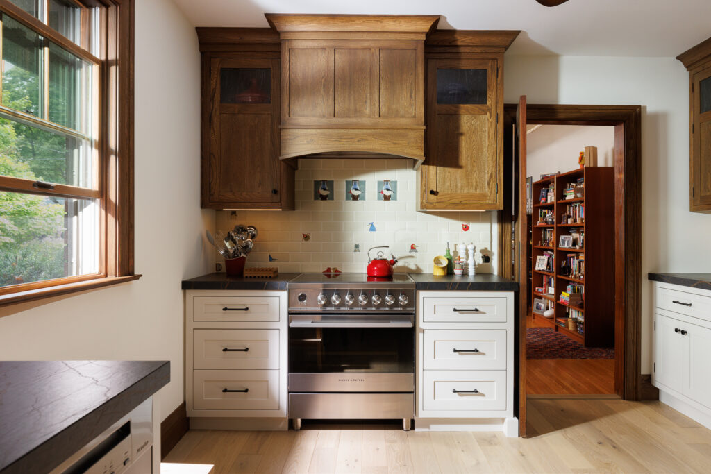 A bright kitchen with wooden and white cabinetry, a stainless steel stove, black countertops, and a red kettle. A window lets in natural light, and an open door reveals a room with bookshelves.