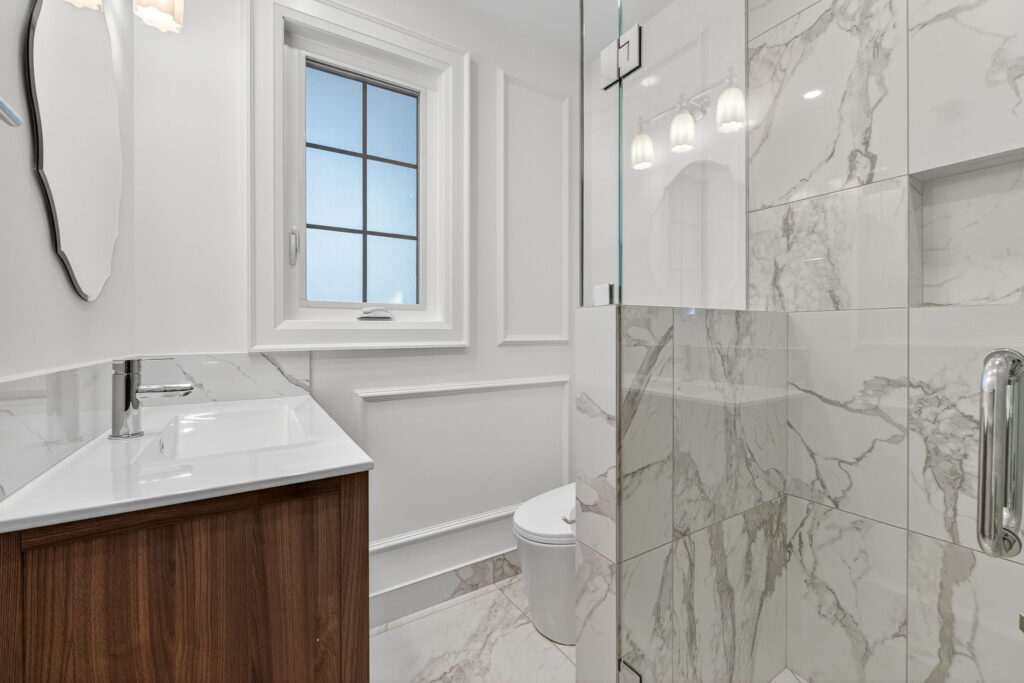 Modern bathroom with white marble tiles, glass shower, wood vanity with sink, wall mirror, and frosted window. Bright lighting and clean, minimalist design.